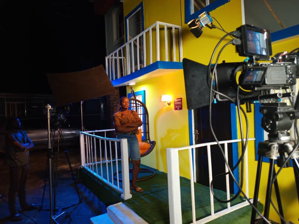 A night shoot on the yellow house set — an actress on the porch, framed on the camera monitor in the foreground