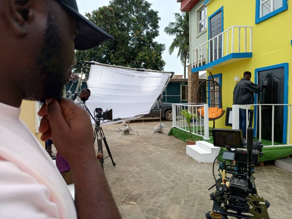 An outdoor shoot at the yellow house set — camera rig and diffusion frame set up while the crew prepares the door